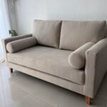 Beige fabric loveseat with bolster pillows on a light tiled floor.
