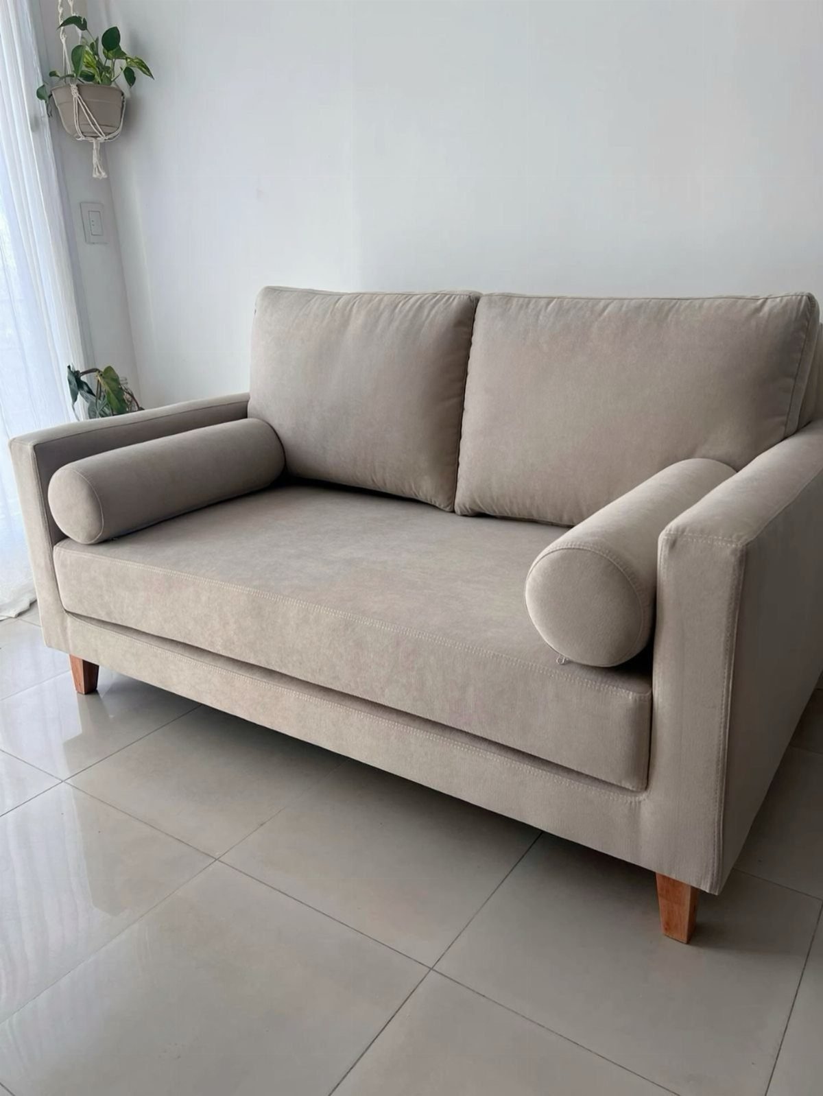 Beige fabric loveseat with bolster pillows on a light tiled floor.