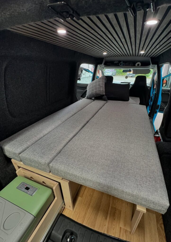 Custom wooden bed frame with grey cushions inside a modern camper van interior.