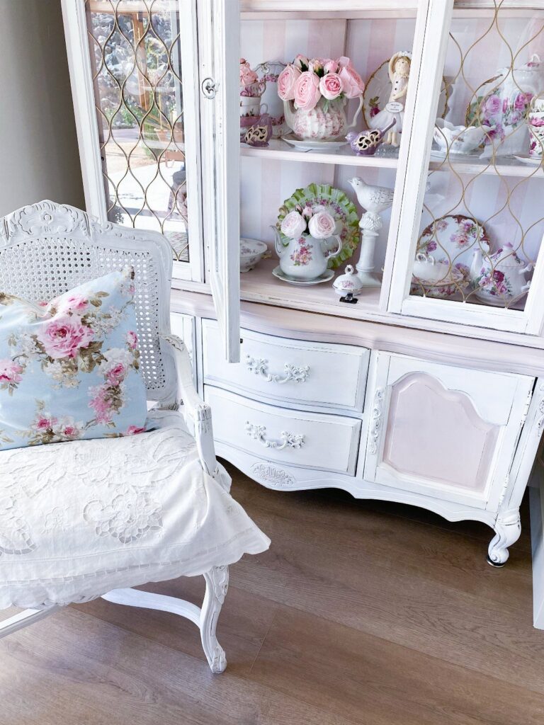 White painted vintage hutch and ornate chair with floral pillow in a room.