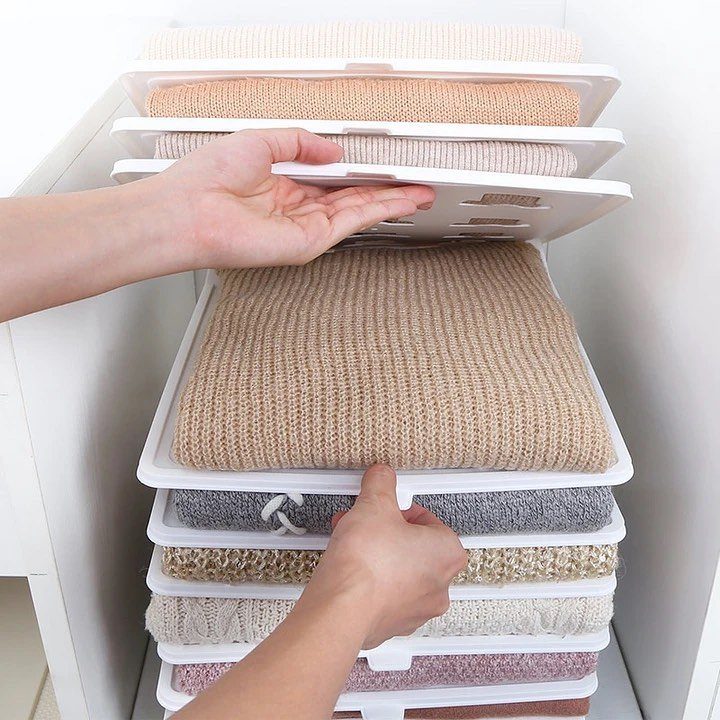 Folded knit sweaters organized in white plastic stacking trays inside a white shelf.